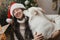 Cute dog playing with owner at stylish christmas tree. Pet and winter holidays. Happy woman in santa hat hugging funny white