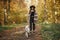 Cute dog walking on background of stylish woman traveler owner in sunny autumn woods. Traveling with pet, loyal companion. Young