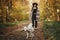 Cute dog walking on background of stylish woman traveler owner in sunny autumn woods. Traveling with pet, loyal companion. Young