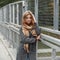 Cute European young woman in a gray elegant stylish coat in a beige vintage checkered scarf posing standing on a bridge