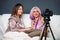 Cute, fashionable girlfriends pose for the camera on a couch with pillows. Modern blogging, photo camera in studio