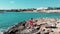 Cute female in red dress walking on rocky beach pier alone enjoying sun and hot wind. Waves colliding with rocks and sun reflects