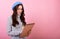 A cute French lady student in a fashionable beret hat with a pen and a notebook in her hands smiles and takes notes on a pink