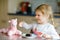 Cute funny toddler girl eating chocolate ice cream at home. Happy healthy baby child feeding plush lama toy with sweet