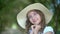 Cute girl portrait in white hat holding hands near face in green garden. Adorable young in summer hat girl smiling and