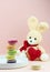 Cute handmade teddy rabbit holding a red heart sitting behinde a white plate with macaroons in trendy pastel colors