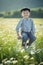 Cute handsome boy wearing jeans and shirt sitting on wooden chair on meadow of chamomiles daisy and smiling enjoying summer day in