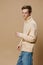 a cute, handsome guy in a long, textured, beige sweater looks at the camera, posing against a plain background. Studio