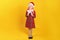 Cute happy little girl holding a box with a christmas gift. Full-length child in a red dress and santa hat on a yellow background