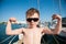 Cute healthy small caucasian boy in sunglasses showing biceps muscle on sea port with yachts on pier