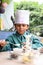 Cute Indian chef boy preparing sundae dish as a part of non fire cooking which includes vanilla ice cream, brownie, coco powder,