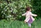 Cute kid girl in a light pink coat and dress, with a ponytail, darts around herself near a blossoming apple tree in spring