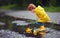 Cute little baby boy launching paper boats in spring puddles, wearing raincoat and rubber boots