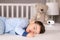 Cute little baby boy in light blue pajamas sleeping peacefully on bed at home with baby monitor camera and soft teddy bear toy at