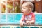 A cute little baby holds to the edge of the tropical pool and looks to the left. Infant girl swim at summer vacation