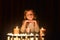 The cute little blonde girl keeps her hands under the chin. Lots of candles are around her, over dark background