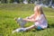 Cute little blonde girl sitting on green grass and putting on roller skates - leisure, childhood, outdoor games and sport concept.