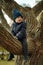 A cute little boy in a blue warm jacket and knit beanie sitting on the branch of big tree. He is afraid to fall down. Image with s