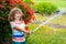 Cute little boy watering flowers in the garden at summer day. Child using garden hose. Funny kid watering plants in the