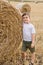 A cute little boy in a white t-shirt and khaki shorts is standing near round bales of hay. Photo session in the field