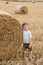 A cute little boy in a white t-shirt and khaki shorts is standing near round bales of hay. Photo session in the field