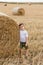 A cute little boy in a white t-shirt and khaki shorts is standing near round bales of hay. Photo session in the field