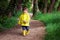 Cute little boy in yellow raincoat and rubber boots holding rhino toy with scared face walking alone lost in dark green forest.