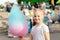Cute little caucasian blond kid girl holding in hand stick with Bright big multicolored cotton candy sweet at city park during