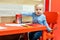 Cute little caucasian blond toddler boy sitting at table and drawing at children area at retail clothes store. Baby spending time