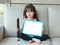 Cute little child girl holding empty white blackboard with big pencil sitting on fabric sofa in library room. Education concept