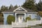 Cute little cottage with blue siding and victorian touches and wood shingles and white picket fence against tall pine trees