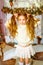 Cute little girl with blond long curly hair in a light knitted sweater by the festive fireplace with garlands