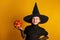 Cute little girl dressed in a witch costume holds a pumpkin jack lantern on a yellow background. Halloween celebration