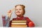 A cute little girl with glasses is sitting at the table, holding a ruler in her hand. Schoolgirl is surrounded by books.