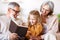 Cute little girl granddaughter smiling when reading book with senior grandparents