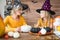 Cute little girl and her mother, both in witch costumes, sitting behind a table in Halloween theme decorated room, smiling.