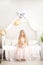 Cute little girl with long blond hair in a dress holds a straw hat sitting on a bed in a white bedroom in the morning. Rustic bedr