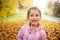 Cute little girl with missing teeth playing with yellow fallen leaves in autumn forest.
