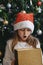 A cute little girl opens a Christmas present with surprise in a New Year`s hat under the tree. Holiday, present, miracle, magic