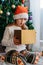 A cute little girl opens a Christmas present with surprise in a New Year`s hat under the tree. Holiday, present, miracle, magic