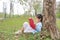 Cute little girl reading book in summer park outdoor lean against tree trunk in the summer garden