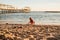 a cute little girl sits on the seashore, playing in the sand and with pebbles.