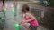 Cute little girl squatting and playing with colored water jets at the fountain in hot summer day.