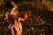 Cute little girl is standing in autumn forest during sunset, holding an orange pumpkin.