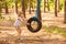 Cute little girl swinging on wheel attached to big tree in forest.