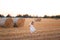 Cute little girl in white dress strolling down a wheat field with big stacks on sunset