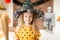 Cute little girl in witch costume with hat is holding a broom and standing in Halloween decorated living room, looking at camera.