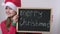 Cute little Santa helper girl shows billboard with text Merry Christmas. Adorable child in red Santa hat holding black