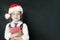 Cute little schoolgirl child in Santa hat with school book on back chalkboard background. Smiling pupil in classroom