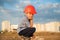 Cute little tired sad kid in orange helmet sitting on background of new buildings and sunset cloudy sky